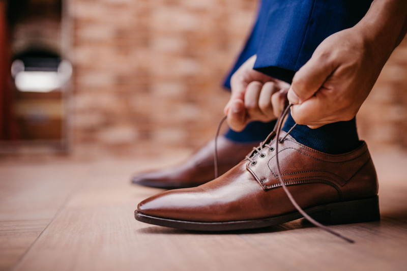 Hands lacing a brown leather derby on a wooden floor.